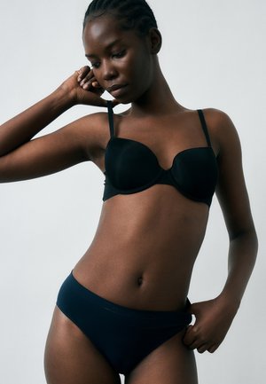 Kvinde iført sort bh og matchende undertøj justerer sin strop, kigger ned med neutralt udtryk mod en ensfarvet baggrund.