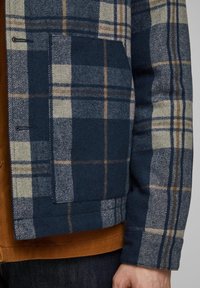 Károvaná vlněná bunda v námořnické modré, krémové a béžové barvě, s náprsní kapsou a knoflíkovým zapínáním. Přes ni je přehozená hořčicová košile.