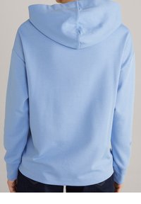 Persona che indossa una felpa leggera blu vista da dietro, con il cappuccio abbassato e pantaloni scuri visibili sotto l'orlo.