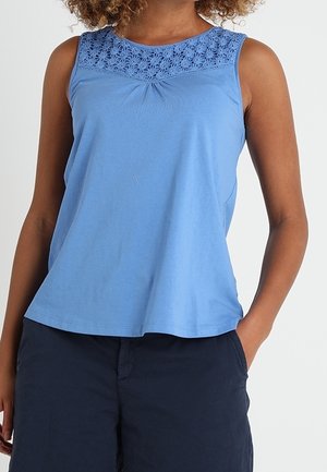 Top sin mangas de color azul claro con detalles de encaje floral en la parte superior del pecho, combinada con pantalones cortos azul marino oscuro, con las manos apoyadas en los bolsillos.