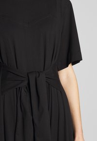 Svart klänning med åtsittande överdel och korta ärmar. Har ett knutet bälte i midjan och plisserad kjol. Mjuk, strukturerad tyg.