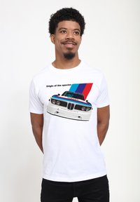 Hvid t-shirt med et grafisk motiv af en klassisk bil med sorte og røde detaljer, blå og røde striber, samt tekst, der siger "Oprindelsen af arten."
