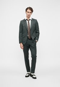 Σκούρο πράσινο ανοιγμένο κοστούμι με λευκό πουκάμισο και καφέ σχέδιο γραβάτα. Τα παπούτσια είναι μαύρα και λευκά loafers. Σχεδίαση στενής εφαρμογής, λεία υφή.