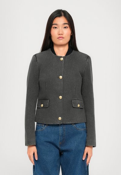 Frau mit langen dunklen Haaren, die eine enganliegende graue Jacke mit goldenen Knöpfen und eine blaue Jeans trägt, steht vor einem hellen, einfarbigen Hintergrund.
