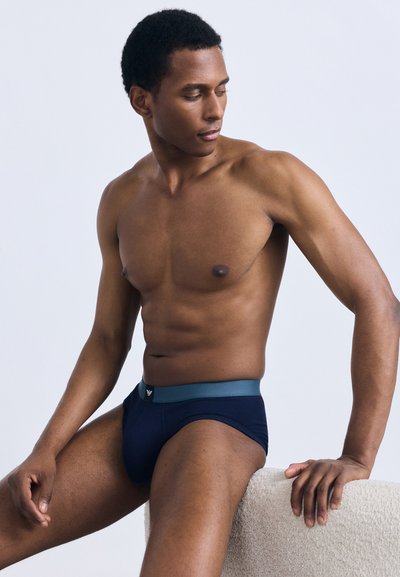 Tamsiai mėlynos trumpikės su kontrastingu pilku liemeniu, pagamintos iš minkštos medžiagos, su logotipo etikete priekyje ir aptempto silueto.