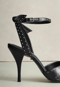 Schwarze High-Heel-Sandale mit glänzender Oberfläche, Nietenakzenten, Fesselriemen mit Schleifendetail und elegantem Plateau-Design.