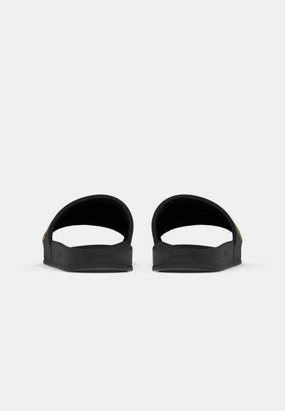 Une paire de sandales noires à enfiler orientées vers l'avant, montrant des sangles courbées et des semelles plates sur un fond blanc uni.