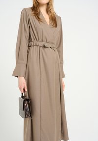 Pitkä, taupe-värinen mekko, jossa on kietaisu-mallinen muotoilu, vyötäröä korostava osa ja hopeiset napit. Pitkät hihat, joissa on levenevät päärmeet. Malli pitää pieniä, tummaa käsilaukkua.