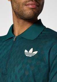 Teal polo-shirt med kvart lynlås og struktureret diamantmønster. Har et hvidt Adidas-logo og klassisk krave-design.