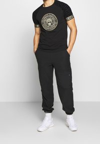 Camiseta negra con gráfico de león dorado y mangas con estampado, combinada con pantalones cargo negros que tienen un bolsillo lateral; usados con zapatillas blancas.