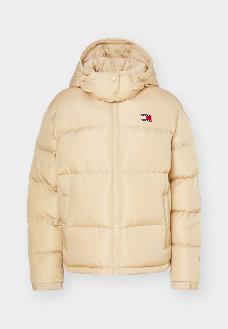 Beige Pufferjacke mit Reißverschluss vorne, elastischen Bündchen und einer Kapuze. Verfügt über ein kleines Logopatch mit rot-weißen und blauen Streifen auf der Brust.
