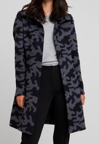 Camouflage jas van een wolmix in grijs en zwart. Voorzien van een reverskraag, knoopsluiting en zijzakken. Gestructureerde pasvorm, tot op de knie.