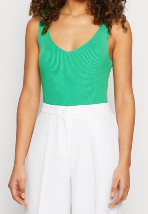 Femme portant un haut vert sans manches à côtes et un pantalon blanc large à taille haute, avec des cheveux bouclés visibles aux épaules.