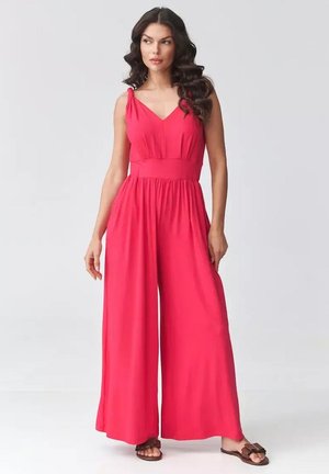 Kobieta o długich falowanych włosach, ubrana w bezrękawnikowy, czerwony, szerokie spodnie kombinezon oraz brązowe sandały, stojąca na tle jasnego, gładkiego tła.