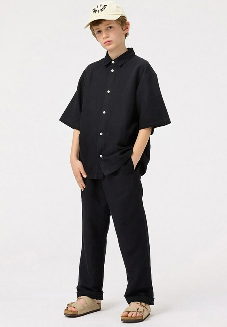 Zwarte korte mouwen overhemd met knopen en bijpassende broek, gecombineerd met beige sandalen en een lichte pet met een bedrukt ontwerp.