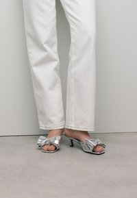 Zilveren metalen hoge hak sandaaltjes met een strikdetail, open teen en een slank ontwerp, gecombineerd met witte rechte broek.