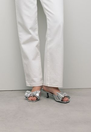 Zilveren metalen hoge hak sandaaltjes met een strikdetail, open teen en een slank ontwerp, gecombineerd met witte rechte broek.
