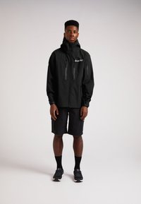 Sort vandtæt jakke med høj krave, to lynlås brystlommer og et logo. Båret med sorte shorts og sorte sportsko.