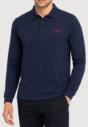 Mann trägt ein dunkelblaues Poloshirt mit langen Ärmeln und rotem "GISADA"-Logo auf der Brust sowie schwarze Hose, eine Hand in der Tasche.