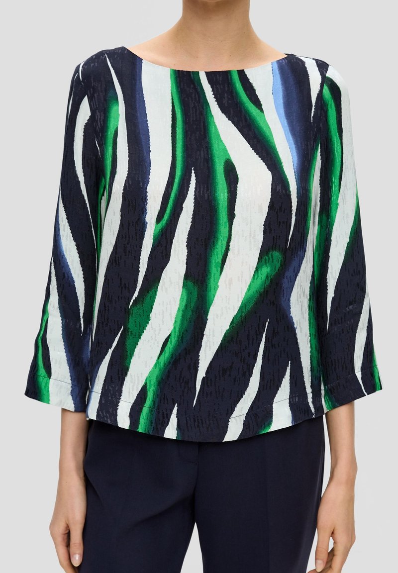 Femme portant un chemisier à manches longues avec un motif rayé abstrait vert, blanc et bleu et un pantalon foncé sur un fond uni.