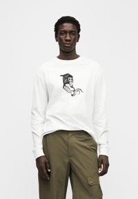 Λευκό μακρυμάνικο T-shirt με μαύρο γραφικό σχέδιο κρανίου με ένα φίδι και κείμενο. Συνδυάζεται με πράσινο cargo παντελόνι που έχει πλαϊνές τσέπες και κουμπιά.