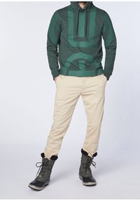 Felpa verde con cappuccio e stampa geometrica, pantaloni beige e stivali impermeabili verde scuro con bordo in pelliccia e dettagli con lacci.