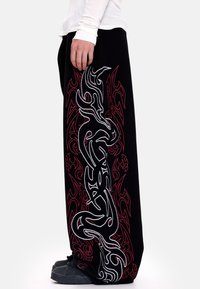 Juodos kelnės su išraiškingais raudonais ir baltais genčių raštais kairėje pusėje, pagamintos iš minkštos medžiagos, laisvo kirpimo.
