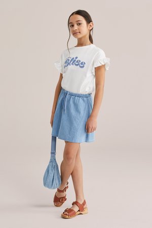 Junges Mädchen trägt ein weißes Shirt mit Rüschenärmeln und dem Schriftzug "Bliss", einen hellblauen Rock, braune Sandalen und hält eine passende hellblaue Tasche.