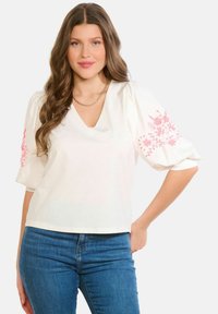 Balta blūze ar V veida izgriezumu un pūstām piedurkn ēm ar rozā ziedu izšuvumiem. Izgatavota no viegla auduma ar brīvu piegriezumu.