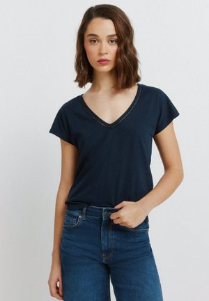 Kobieta o brązowych włosach do ramion, ubrana w granatowy t-shirt z dekoltem w serek i niebieskie jeansy, stojąca na jednolitym jasnym tle.