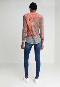 Multicolored blouse with floral and geometric patterns, semi-transparent material, long sleeves, paired with blue skinny jeans and white sneakers.