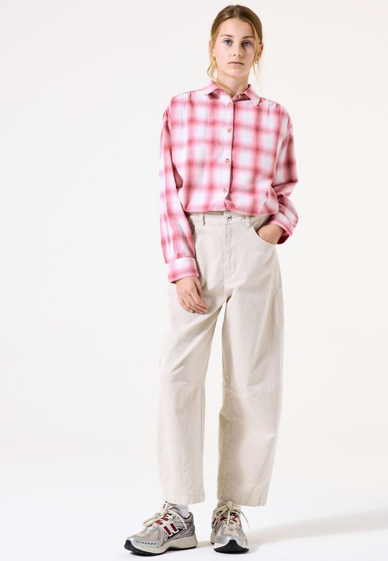 Rosa karierte Hemd mit langen Ärmeln, kombiniert mit beigen weit geschnittenen Hosen und mehrfarbigen Sportschuhen. Schlichter Hintergrund.