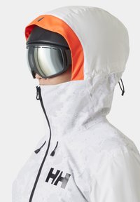 Hvit skijakke med oransje hette, med svart logo, høy krage og glidelås. Inkluderer svarte skibriller med reflekterende linser.