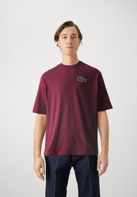 T-shirt in cotone bordeaux con scollo rotondo e maniche corte. Presenta un logo di coccodrillo verde ricamato sul lato sinistro del petto.