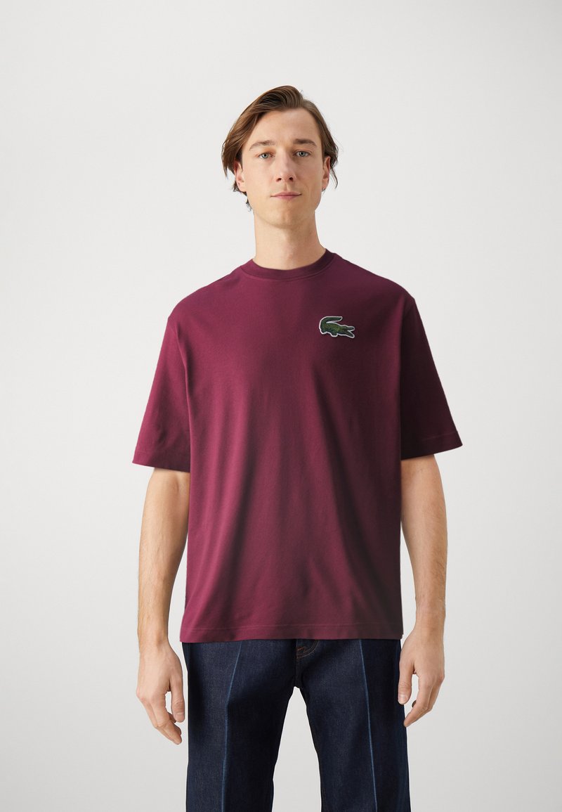 T-shirt in cotone bordeaux con scollo rotondo e maniche corte. Presenta un logo di coccodrillo verde ricamato sul lato sinistro del petto.