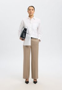 Camicia bianca a maniche lunghe con taschino sul petto, pantaloni beige su misura, che tiene una borsa nera e indossa décolleté nere appuntite. Sfondo neutro.