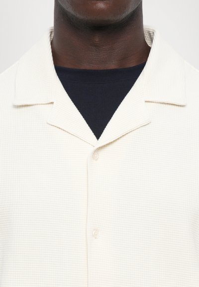 Weißes, strukturiertes Button-up-Hemd mit Kragen. Über einem marineblauen Rundhalshemd getragen, das Details des Stoffes und der Nähte zeigt.