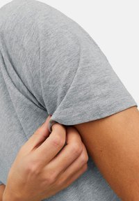 Maglietta grigia a maniche corte con una texture morbida. Una mano tira la manica, mostrando la cucitura e il sottile motivo del tessuto.
