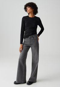 Vrouw met krullend haar die een zwart shirt met lange mouwen, grijze wijde jeans en zwarte schoenen draagt, staande met één hand op de heup.