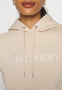 Béžová mikina s kapucí vyrobená z měkké tkaniny. Obsahuje velké bílé logo "Calvin Klein", šňůrky s kovovými konci ve stříbrné barvě a klokankovou kapsu.