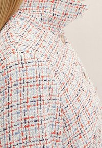 Veste en tweed avec un motif tissé texturé composé de fils blancs, rouges et bleus. Col montant et détail de boutons sur le côté.