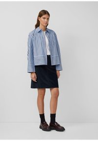 Gestreepte blauwe en witte jas met een kraag, gecombineerd met een marineblauwe rok. Model draagt donkere sokken en bruine schoenen met een stevige zool.