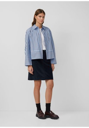 Veste rayée bleue et blanche avec un col, associée à une jupe marine. La mannequin porte des chaussettes foncées et des chaussures marron à semelle épaisse.