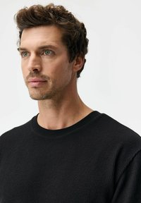 Czarny dzianinowy sweter z okrągłym dekoltem i teksturowaną tkaniną, prezentowany na męskim torsie z krótkimi, kręconymi włosami na jasnym tle.