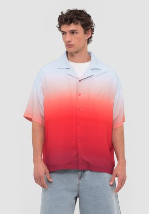 Man die een kortemouwen overhemd met knopen draagt met een verloop van lichtblauw naar rood en lichtblauwe spijkerbroek, staand tegen een effen achtergrond.