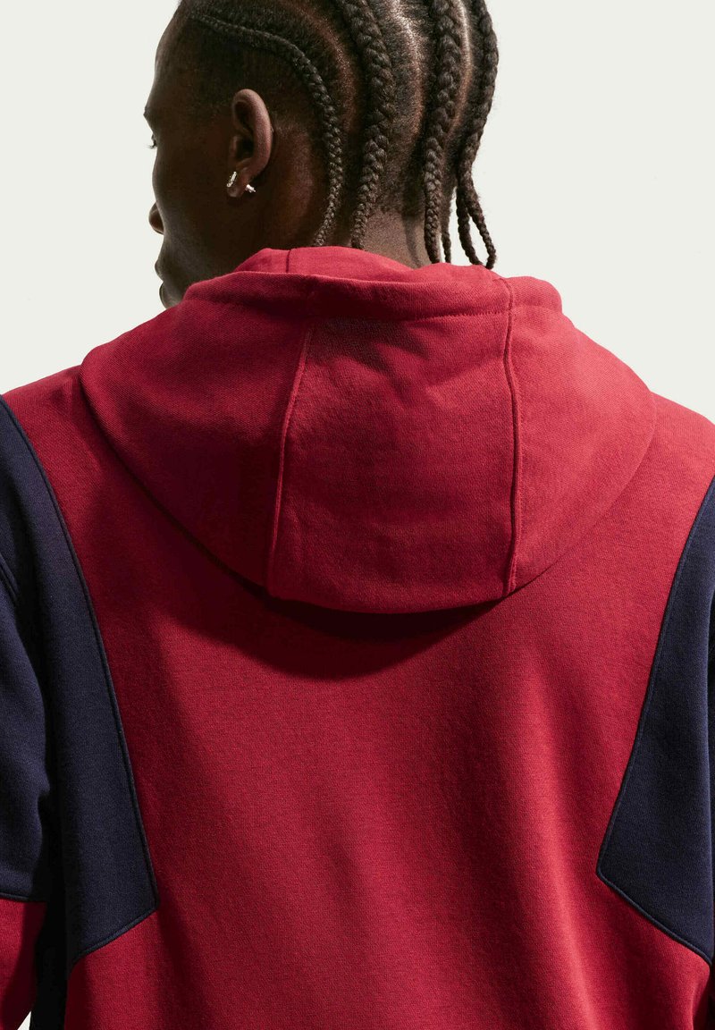 Persona con i capelli intrecciati che indossa una felpa rossa con maniche blu scuro, vista da dietro su uno sfondo chiaro e uniforme.