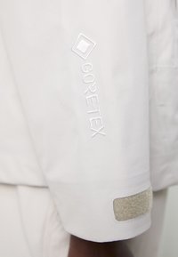 Vit vattentät jacka med broderad "GORE-TEX"-logotyp på ärmen, med en texturerad Velcro-justering vid ärmslutet. Slät tygfinish.