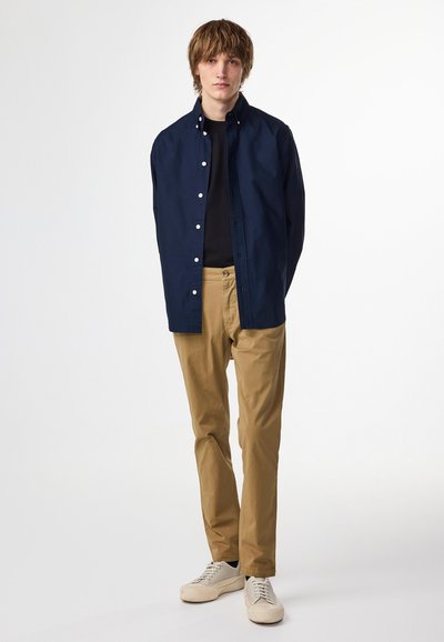 Camicia di cotone blu navy con bottoni bianchi, t-shirt nera sotto, pantaloni khaki e sneakers beige. Il modello è in piedi contro uno sfondo semplice.
