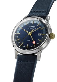 Die Timex Marlin Uhr verfügt über ein rundes Gehäuse aus Edelstahl, ein blaues, mehrtexturiertes Zifferblatt, goldene Stundenmarkierungen, einen roten GMT-Zeiger und ein blaues Lederarmband.