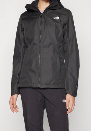 Person, der bærer en sort lynlåstrøje med hætte og matchende bukser, begge med The North Face-logo på brystet og låret.